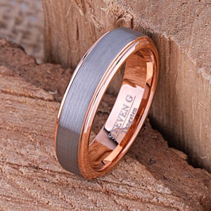 May include: A close-up of a tungsten ring with a brushed grey centre and rose gold edges. The ring is engraved with the text "STEVEN G Tungsten". The ring is set against a wooden background.