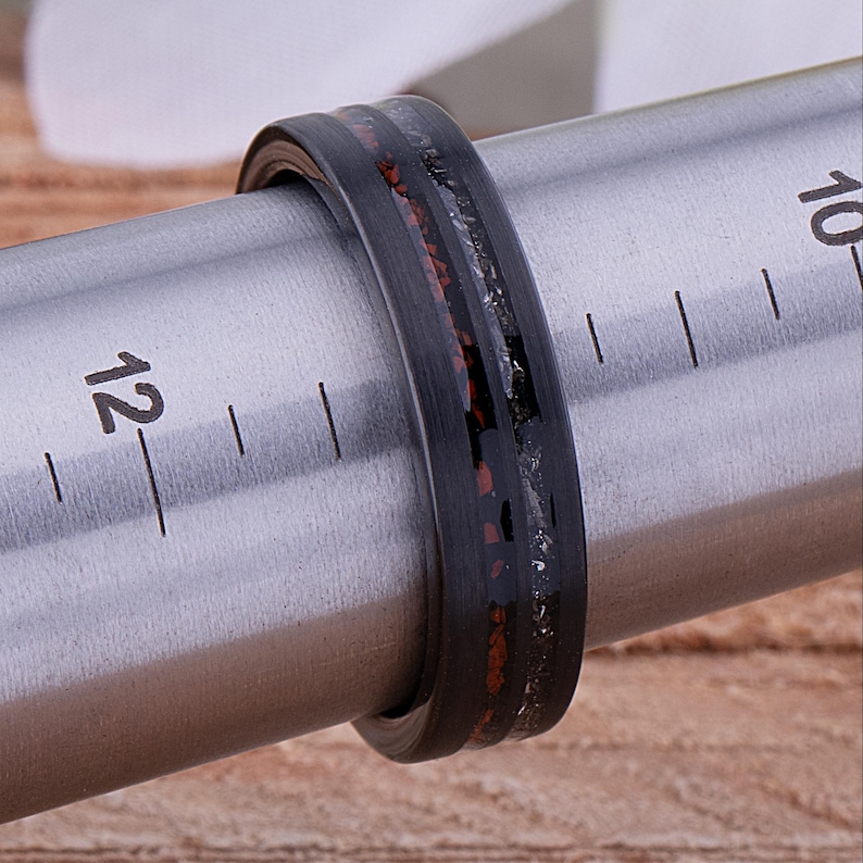 May include: A black and silver ring with a red and black inlay. The ring is being measured on a metal ruler.