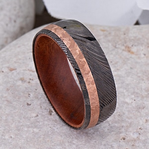 May include: A black and grey Damascus steel ring with a rose gold inlay and a brown wooden band.