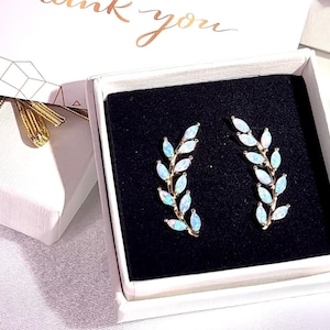 May include: A pair of gold earrings with opal stones in the shape of leaves. The earrings are in a white box with a black interior. The box has a thank you note on top.