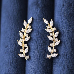Può includere: Un paio di orecchini a lobo allungati color oro con un design a forma di ramo, decorati con pietre trasparenti a taglio marquise e rotondo. Gli orecchini sono disposti in un orientamento verticale e curvo, con una montatura metallica incastonata.