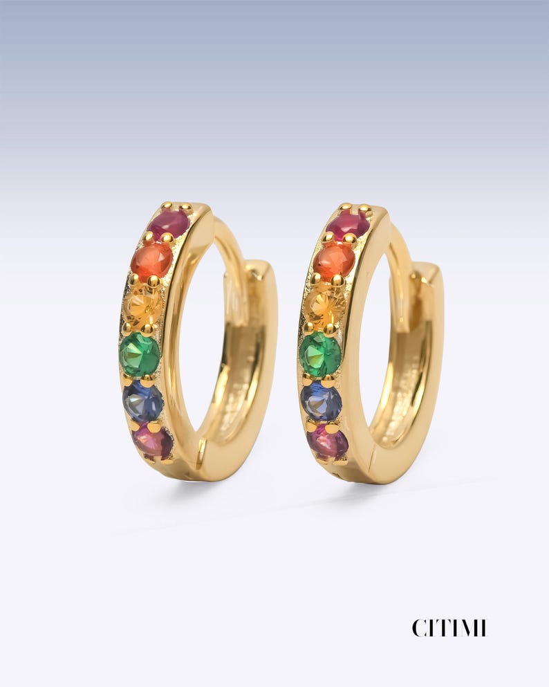 Pu&ograve; includere: Un paio di orecchini a cerchio dorati con una fila di piccole pietre preziose rotonde e multicolori nei toni del rosso, arancione, giallo, verde, blu e viola. La parola CITIMI &egrave; stampata in nero nell'angolo in basso a destra.