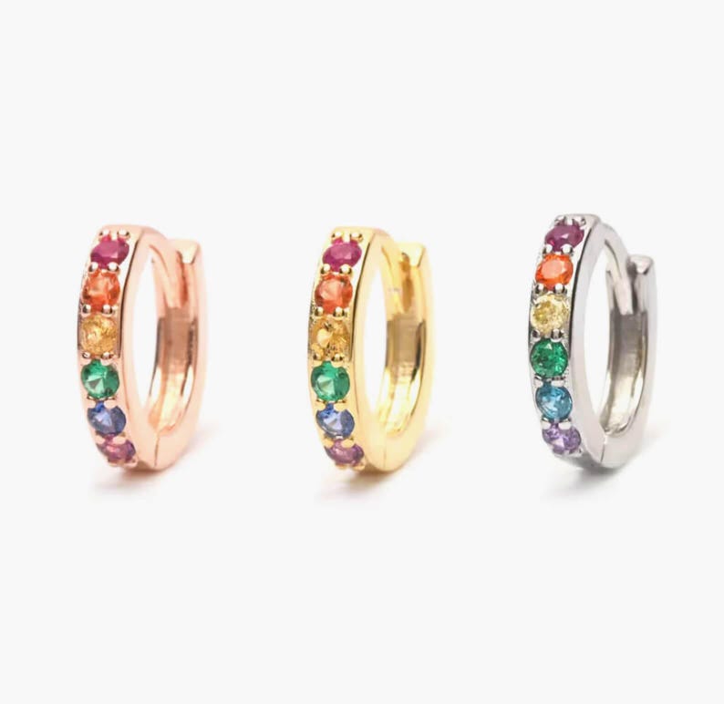 May include: Three pairs of rainbow hoop earrings. The earrings are made of silver, gold, and rose gold. Each earring has a row of colourful gemstones.
