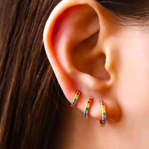 May include: Three gold hoop earrings with rainbow coloured gemstones. The earrings are small and delicate.