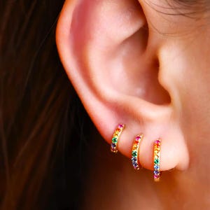 May include: Three gold hoop earrings with a rainbow of colourful gemstones set in the band. The earrings are shown in a person's ear.