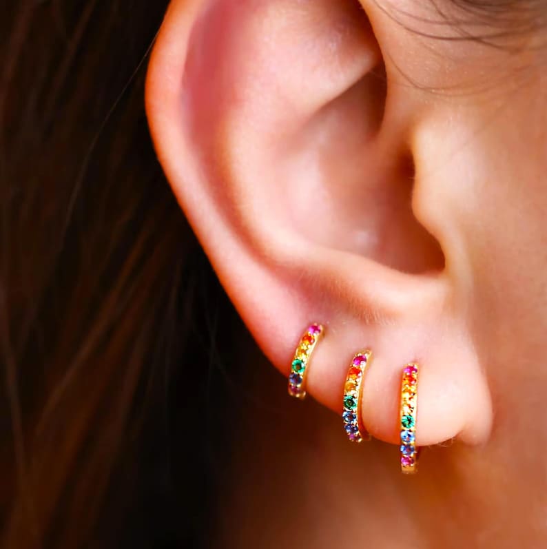 May include: Three gold hoop earrings with a rainbow of colourful gemstones set in the band. The earrings are shown in a person's ear.