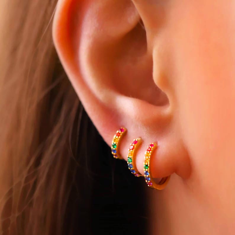 May include: Three gold hoop earrings with rainbow coloured cubic zirconia stones. The earrings are worn in the earlobe.