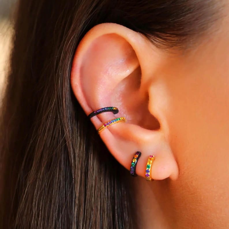 May include: Two gold ear cuffs with a rainbow of small gemstones set in black metal. The ear cuffs are worn on the upper part of the ear.