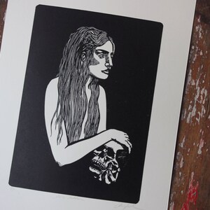 Puede incluir: Grabado en lin&oacute;leo en blanco y negro que representa a una mujer con pelo largo sosteniendo una calavera. La obra de arte est&aacute; sobre un fondo negro, con la mujer y la calavera en blanco. La impresi&oacute;n est&aacute; sobre un fondo blanco.