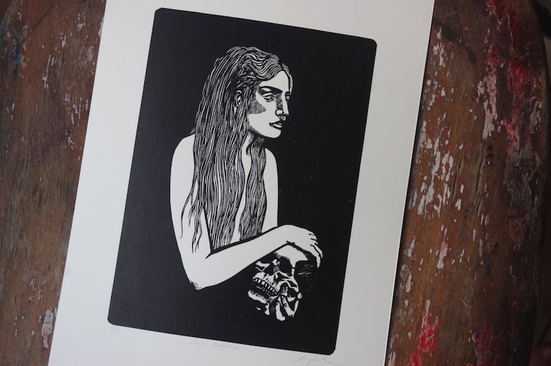 Puede incluir: Grabado en lin&oacute;leo en blanco y negro que representa a una mujer con pelo largo sosteniendo una calavera. La obra de arte est&aacute; sobre un fondo negro, con la mujer y la calavera en blanco. La impresi&oacute;n est&aacute; sobre un fondo blanco.