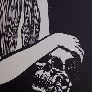Puede incluir: Grabado en lin&oacute;leo en blanco y negro que presenta una calavera y una figura con pelo largo. La obra de arte tiene un dise&ntilde;o de alto contraste, con la calavera en primer plano y la forma de la figura en el fondo. El estilo recuerda a las xilograf&iacute;as.