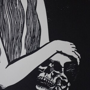 Puede incluir: Grabado en lin&oacute;leo en blanco y negro que representa una calavera sostenida por una persona. Se ven el brazo y el pelo largo de la persona, con los detalles y los dientes de la calavera claramente definidos. La obra tiene un alto contraste y un tema g&oacute;tico.