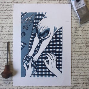 Peut inclure: Une estampe bleue et blanche représentant un chien interagissant avec des mains, avec des outils de couture en arrière-plan. Un outil de sculpture sur bois et un tube de peinture noire sont également présents. L'estampe est sur fond blanc avec une écriture vintage.