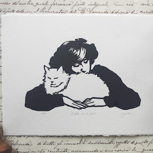 Op de afbeelding: Een zwart-wit linosnede van een vrouw met kort haar die een witte kat vasthoudt. De vrouw draagt een donker shirt en de kat ligt in haar armen. De print is getiteld "Ella & El Gato".
