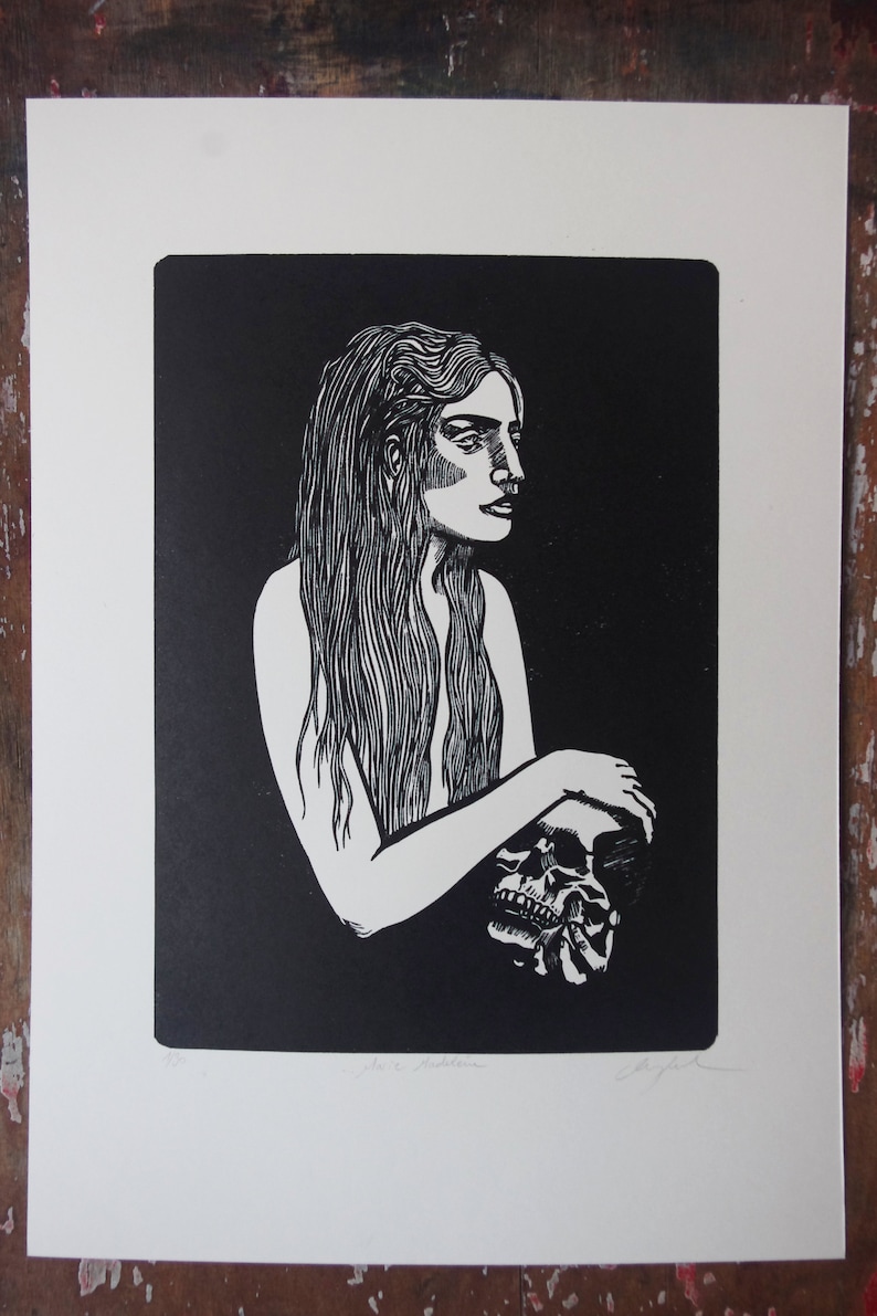 Puede incluir: Grabado en blanco y negro que representa a una mujer con pelo largo sosteniendo una calavera. La obra de arte, de estilo lin&oacute;leo, tiene un fondo oscuro y est&aacute; impresa en papel blanco. La imagen est&aacute; firmada en la esquina inferior derecha.