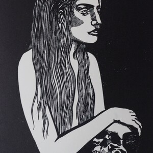 Puede incluir: Grabado en lin&oacute;leo en blanco y negro de una mujer con cabello largo sosteniendo una calavera. La imagen de alto contraste presenta un fondo oscuro y una figura blanca, con un trabajo de l&iacute;neas detallado, evocando un tema g&oacute;tico.