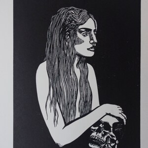 Puede incluir: Grabado en lin&oacute;leo en blanco y negro de una mujer con pelo largo sosteniendo una calavera. La obra de arte muestra un perfil lateral de la mujer, con la calavera en sus brazos. La impresi&oacute;n est&aacute; sobre papel blanco, con la firma del artista.