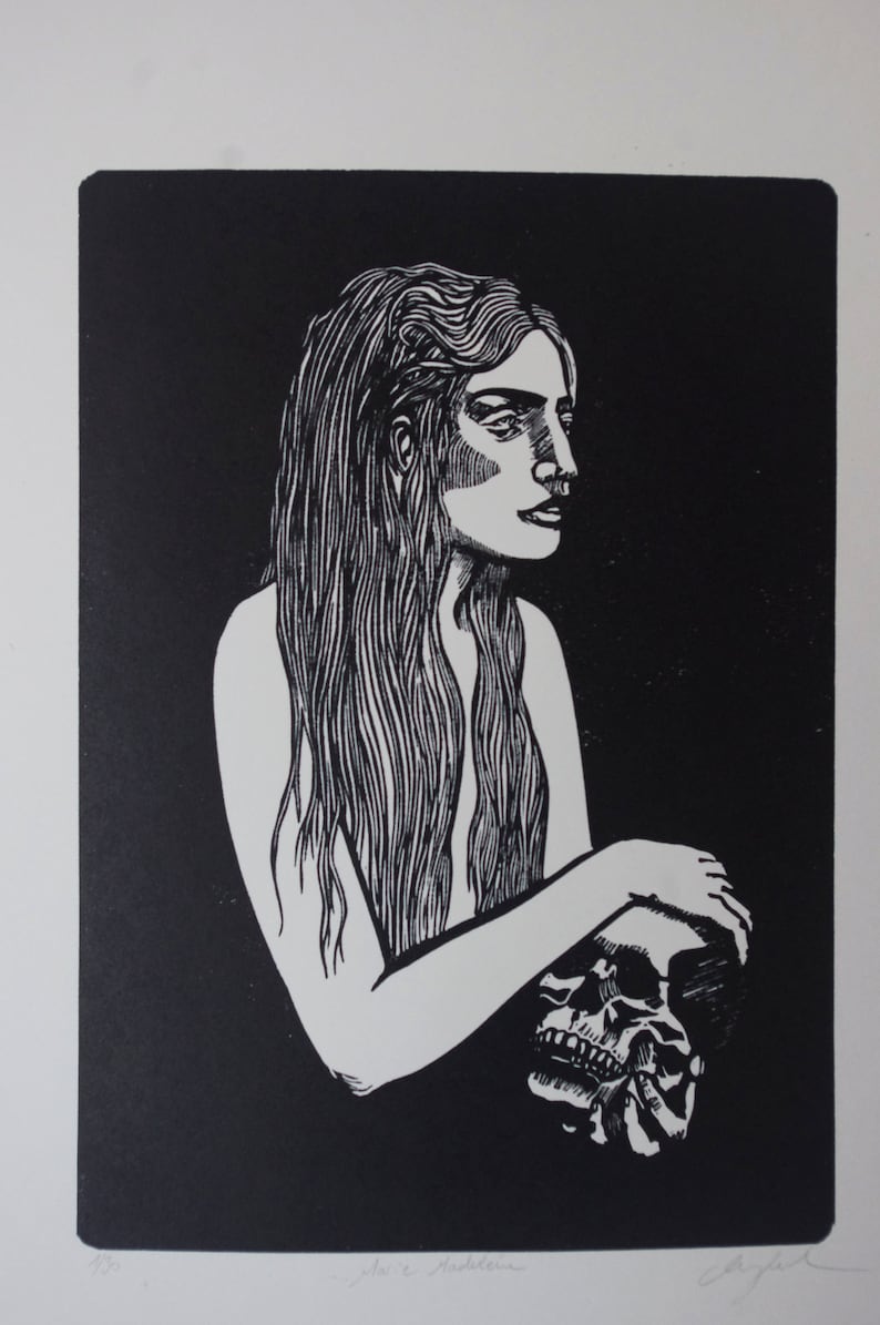 Puede incluir: Grabado en lin&oacute;leo en blanco y negro de una mujer con pelo largo sosteniendo una calavera. La obra de arte muestra un perfil lateral de la mujer, con la calavera en sus brazos. La impresi&oacute;n est&aacute; sobre papel blanco, con la firma del artista.