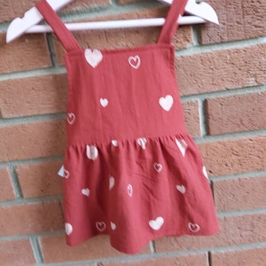 A  baby pinafore dress 9-12 months in rustic red cotton with hearts, it has adjustable shoulder straps, gathered skirt & wooden buttons