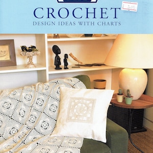 May include: A cream-coloured crochet blanket and cushion with a geometric pattern on a green sofa. The image also shows a book titled "CROCHET DESIGN IDEAS WITH CHARTS" by DMC, along with decorative items on a shelf and a side table.