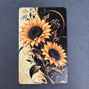 May include: A rectangular card featuring a vibrant illustration of two sunflowers with golden-yellow petals and dark centers. The design incorporates black foliage, delicate sprigs of baby's breath, and thin golden circular lines against a split background.