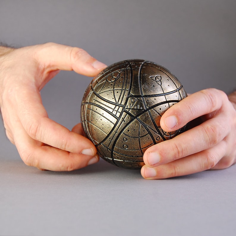 May include: A gold and black sphere with intricate geometric patterns. The sphere is being held in two hands.