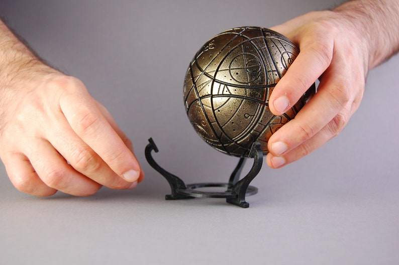 May include: A gold-colored sphere with intricate patterns and lines, resting on a black metal stand with three legs. The sphere is being held by a person's hand.