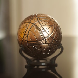 Peut inclure: Une sphère puzzle de couleur bronze avec des motifs géométriques et des lignes complexes. Elle est posée sur un socle noir orné. La surface de la sphère présente une texture avec de petites indentations circulaires.