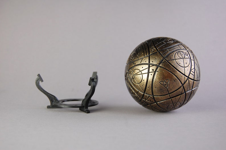 May include: A gold-colored sphere with intricate patterns and a black metal stand. The sphere has a textured surface and appears to be made of metal.