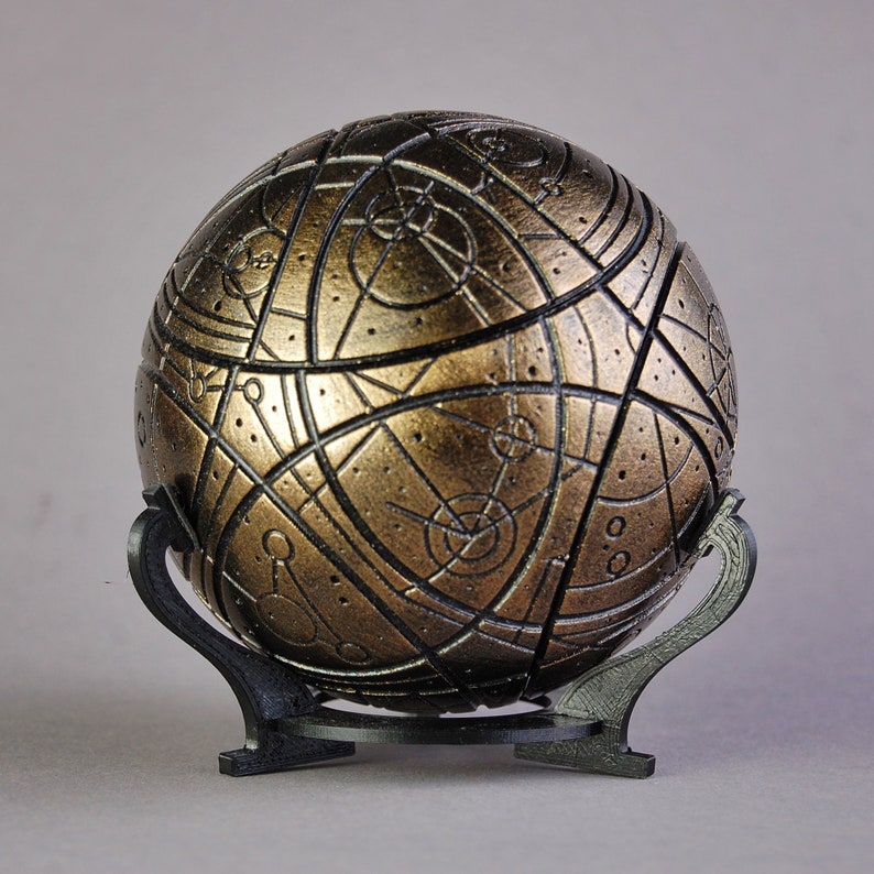 May include: A gold-colored sphere with intricate black markings and a black stand. The sphere has a textured surface and appears to be made of metal.