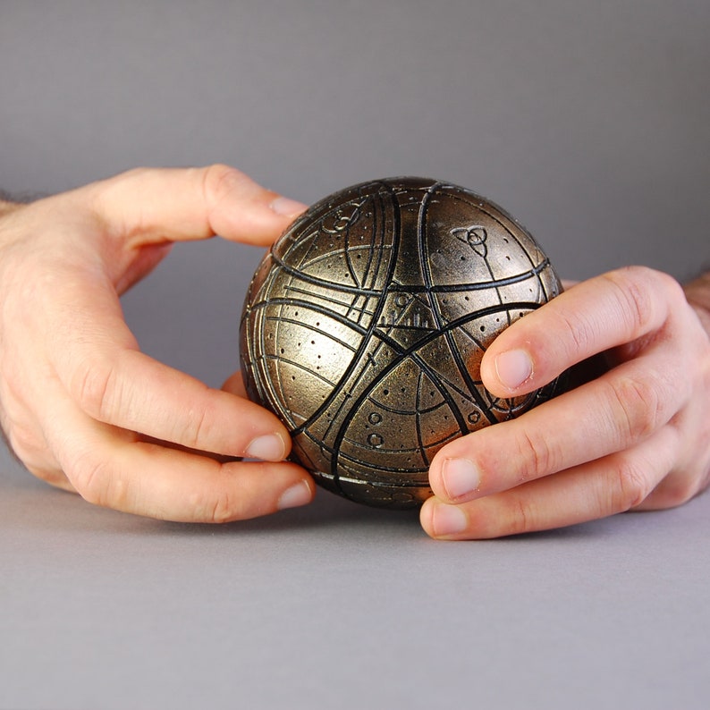 May include: A gold and black sphere with intricate geometric patterns and lines. The sphere is being held by two hands.