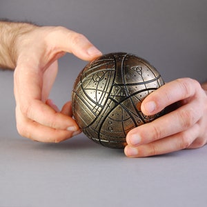 May include: A gold-colored sphere with intricate geometric patterns and lines. The sphere is being held by two hands.