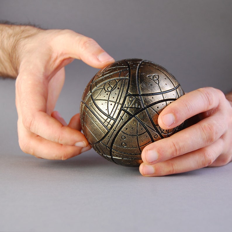 May include: A gold-colored sphere with intricate geometric patterns and lines. The sphere is being held by two hands.