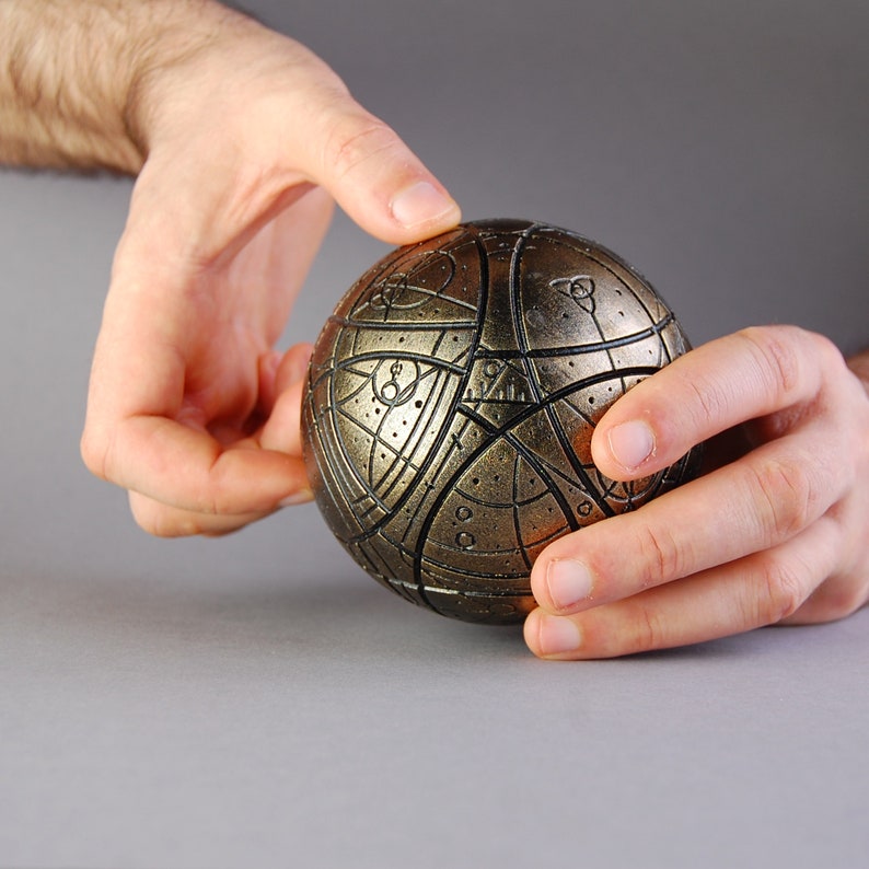 May include: A gold-colored sphere with intricate patterns and lines, held in two hands.