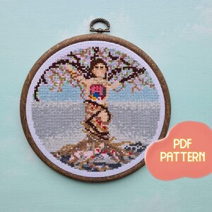 May include: A cross-stitch pattern of a tree with a woman's body inside the trunk. The woman's body is made up of different colored circles and shapes. The tree has branches with leaves and roots. The pattern is in a wooden embroidery hoop. The text "PDF PATTERN" is in a pink cloud shape.
