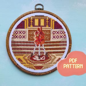 May include: A cross-stitch pattern of a colorful, abstract design in a wooden embroidery hoop. The pattern is in shades of red, orange, yellow, and brown. The hoop is on a light blue background. The text "PDF PATTERN" is in a pink cloud shape.