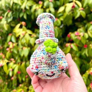 May include: A crocheted amigurumi in the shape of a bong with a green clover on top. The bong is made with a variety of colorful yarns and has a white base with black eyes.
