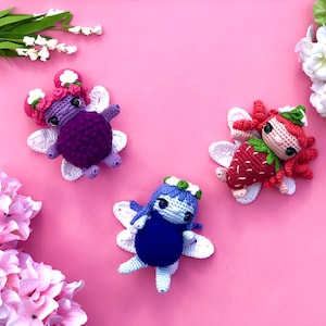 May include: Three crocheted fairy dolls with wings. The fairies are dressed in purple, red, and blue. The fairy on the left is wearing a purple dress with white wings. The fairy in the middle is wearing a blue dress with white wings. The fairy on the right is wearing a red dress with white wings.