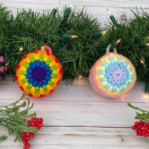 May include: Four crocheted Christmas baubles in a rainbow colour scheme. The baubles are round and have a starburst pattern in the centre. They are hanging from a green garland with white lights.