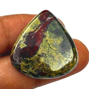 May include: A teardrop-shaped polished gemstone. The stone displays a blend of colours, including deep red, olive green, and gold. The surface is smooth and reflective, with natural patterns and colour variations. The gemstone is held in a hand.
