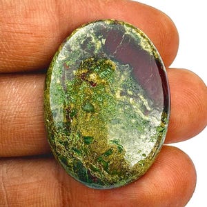 May include: An oval gemstone displaying a blend of green, gold, and burgundy hues. The polished surface reflects light, highlighting the natural patterns and textures. The stone is held in a hand.