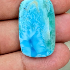 Könnte beinhalten: Ein rechteckiger Cabochon-Edelstein mit polierter Oberfläche, der eine lebendige Mischung aus himmelblauen, türkisfarbenen und weißen Marmormustern aufweist. Der Stein zeigt natürliche Einschlüsse und einen glatten, abgerundeten Rand.