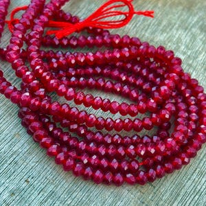 May include: A long strand of small, faceted, deep red beads arranged in multiple loops. The beads have a reflective, polished surface and are strung together on a thin, bright red thread that is tied in a small bow at the top of the arrangement.