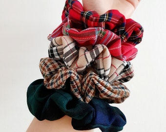 May include: A collection of plaid scrunchies in red, tan, green, and navy blue. The scrunchies are made of fabric and are gathered to create a ruffled effect. The scrunchies are stacked on top of each other.