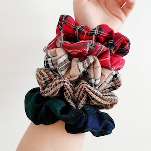 May include: A selection of fabric scrunchies in various tartan patterns, including red, beige, and green. The scrunchies are gathered and layered, showcasing a range of colours and textures. The scrunchies are stacked on top of each other.