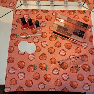 May include: A flat lay featuring beauty products on a pink strawberry-patterned cloth. Includes an eyeshadow palette, nail polish bottles, cotton pads, a cuticle pusher, and eyeglasses. The palette has various shades of eyeshadow.