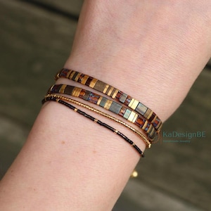 Puede incluir: Un conjunto de tres pulseras superpuestas. Dos pulseras presentan cuentas planas rectangulares en tonos bronce, cobre y gris oscuro metalizados. La tercera pulsera consiste en pequeñas cuentas redondas de color marrón oscuro.