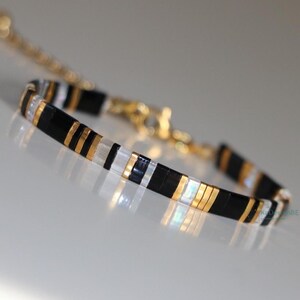 Puede incluir: Una pulsera de cuentas con un patrón repetitivo de cuentas rectangulares en negro, oro metálico y blanco translúcido. La pulsera incluye una cadena extensora de metal dorado y un cierre de mosquetón, con un diseño fino y estructurado.