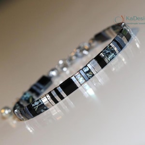 Op de afbeelding: Een armband met een mix van zwarte, zilveren en rechthoekige kralen met patronen. De armband heeft een mix van texturen en kleuren, met een sluiting. De sieraden worden gepresenteerd op een reflecterend oppervlak.