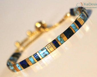 May include: A gold-toned bracelet with alternating rectangular segments of turquoise, navy blue, and gold. The bracelet has a delicate chain and clasp. The handmade jewelry is displayed on a light-colored surface.